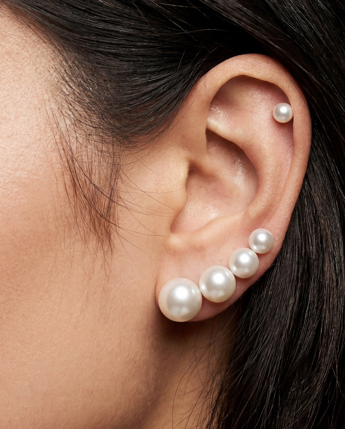 A close-up of a model's ear showcasing a stack of real pearl ear cuffs and stud earrings. The stack features three pearl ear cuffs of varying sizes, with the largest cuff displaying three white pearls of graduated sizes, alongside a single small pearl stud on the opposite helix.